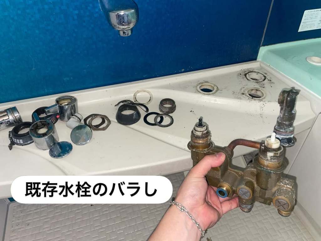 手に持たれた古い水栓のバラシ。周囲には水栓部品と工具が置かれている。